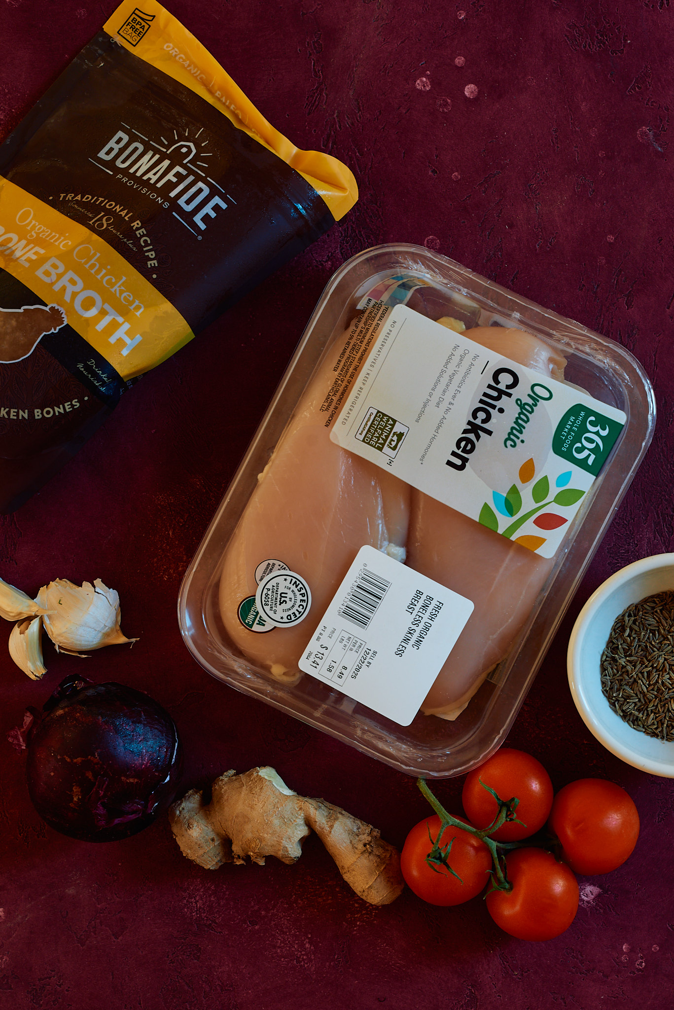 This is a picture of ingredients required for making the indian chicken soup. - chicken, onion, garlic, ginger, cumin seeds, chicken bone broth, tomatoes are shown.